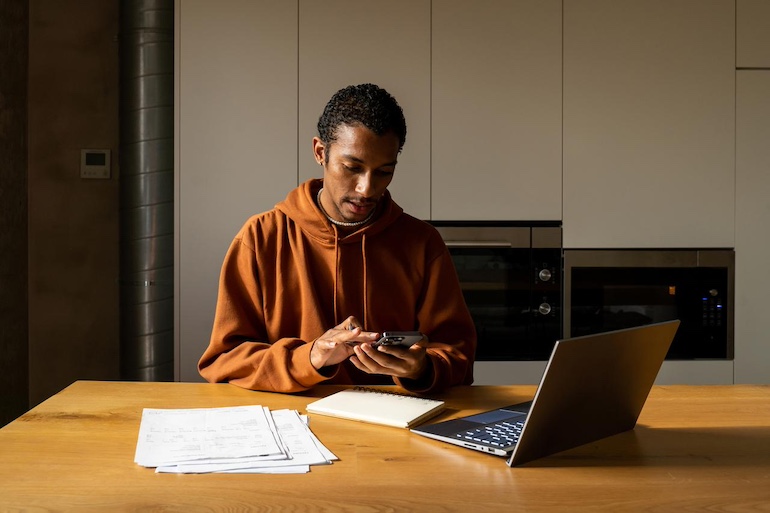 Junger Vermieter sitzt am Tisch vor seinem Laptop, dort liegen einige Formular und er rechnet angestrengt mit seinem Smartphone.